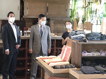 写真:洋服などが並ぶ棚の上に、廃材の数十個におよぶガラス瓶を活用して作られた照明が吊り下げられている。それらを目の前にして、説明を聞く衛藤大臣。