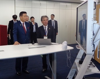 写真:写真左側に、人の上半身より大きなモニターが設置されている。モニター正面に立ち、モニターを見ている衛藤大臣。