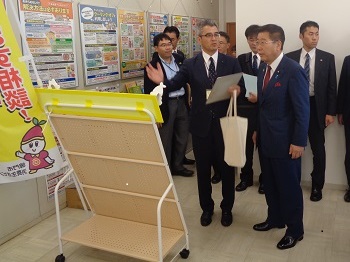写真:消費者トラブルの注意喚起のポスターが並ぶ廊下の先、展示物を見ながら、説明を受ける衛藤大臣。