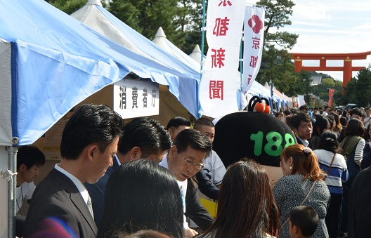 写真:屋外。複数のテントが並ぶ。テント前の通路となっている部分は、人であふれている。消費者庁と書かれたテントの入り口で、チラシの配布を行う衛藤大臣。