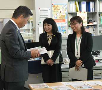 写真:本棚や机が見える執務室、二人の女性職員を前に、衛藤大臣がテキストを手に取り眺めている。