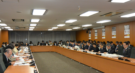 写真:机が大きくコの字型に配置された部屋。20人強が席に付き、その後ろにも椅子が配置され、50人を超える出席者が座っている。