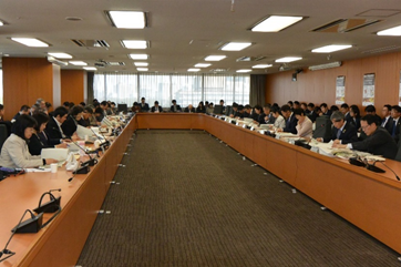 写真:机が大きくコの字型に配置された部屋。20人強が席に付き、その後ろにも椅子が配置され、50人を超える出席者が座っている。