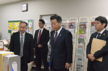 写真:複数の書籍が並ぶ前に立ち、説明を受けている衛藤大臣。