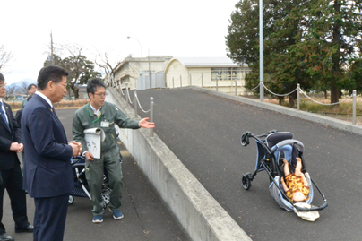 写真:赤ん坊の人形を載せたベビーカーが、坂道で後ろ向きに転倒している。その様子を見ながら説明を受ける衛藤大臣。