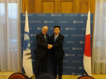 写真:OECDグリア事務総長と衛藤大臣が両手で握手をしている。グリア事務総長の横にはOECDの旗が、衛藤大臣の横には日本国旗が、立てかけられている。