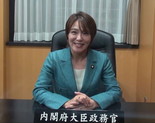 席に着き、カメラに向かって微笑む今井大臣政務官