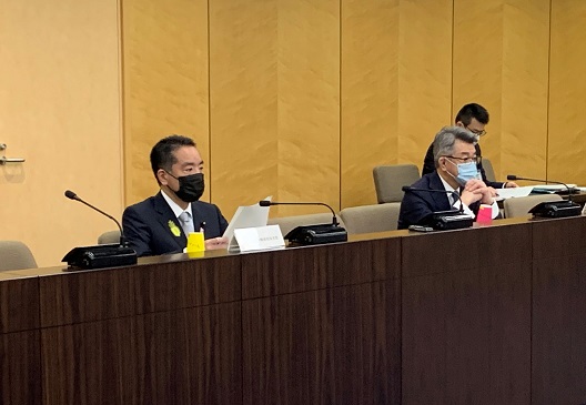 写真:会議用の長い机に着席し、発言する井上大臣。黒いマスクをし、書類を手にしている。スーツの胸元には、「社会を明るくする運動」のシンボルの黄色い羽根を着けている。