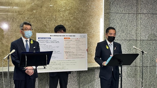 写真:書類を置くための譜面台とスタンドマイクの設置された前に立ち、取材に応じている井上大臣。大臣の後ろでは、職員が「健全な携帯電話市場の実現に向けた取組の推進」のパネルを持って立っている。