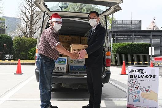 写真:ワゴン車の前で、井上大臣が、赤いサンタ帽を被ったフードバンク団体の職員に災害用備蓄食料の入った箱を渡している。