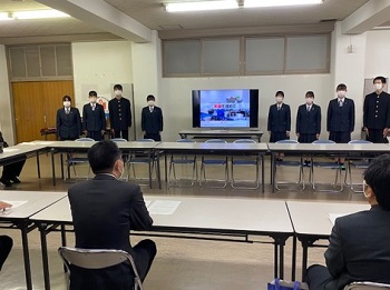 写真:テレビモニターを中心に左右に4人づつ生徒たちが並んでいる。向かい合う机で席につき、井上大臣が説明を聞いている。