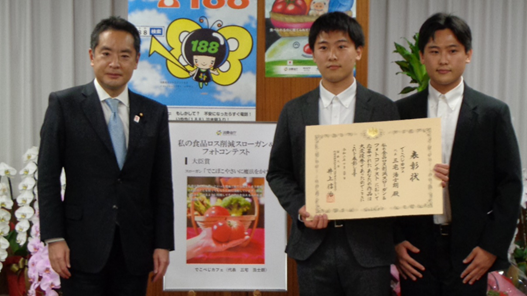 写真:画面右側に井上大臣、左側には賞状を手にした受賞者二人が、受賞作品の写真のボードを後ろに、並んでいる。