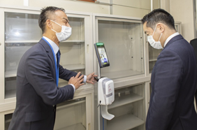 写真:画面中央に設置された消毒ディスペンサーの前に立ち、話を聞く井上大臣。消毒ディスペンサーの左側にはスーツ姿の職員が立ち、大臣に説明をしている。