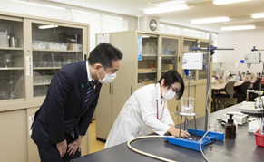 写真:実験机の前で、液体のハンドソープを配合している白衣姿の研究員、その左横で、井上大臣がその様子を見ている