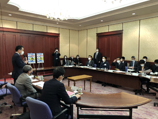 写真:画面左側、机の前には井上大臣、吉村知事、松井市長が横並びに座り、井上大臣が立ち上がって話をしている。向かい合うようにして横に並べられた会議机の前に、複数の参加者が並んで座っている。