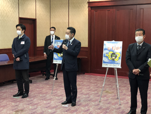 写真:画面中央に井上大臣が立っている、その後ろには、消費者ホットラインの啓発ポスターが飾られている。