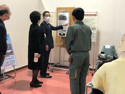 写真:ガス湯沸かし器の実物を前に井上大臣が立ち、職員が横から大臣に説明している
