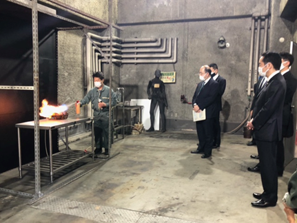 写真:左側に鉄の作業台が置かれ、その上にスプレー缶が置かれている。職員が、噴射剤でスプレー缶に火を噴きかけて、その後ろに少し離れて、井上大臣及び職員がその様子を見ている