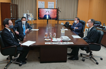 写真:大きな机に、間隔をあけて、4人が座っている。机の奥側にはモニターが置かれており、一人の人物が映し出されている。