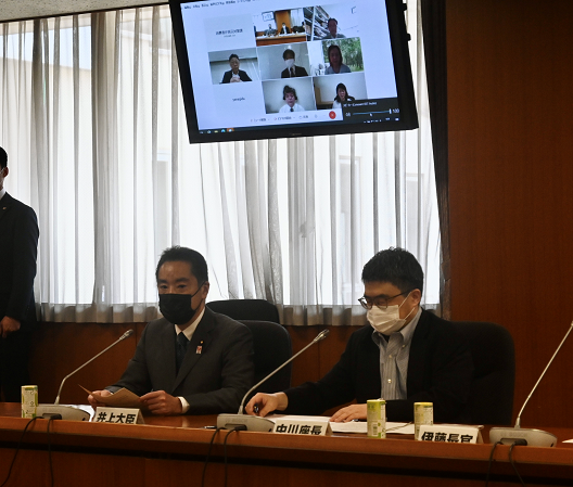写真:中川座長と並んで席に着く井上大臣(写真左側)二人の頭上には、天井からモニターが設置され、オンラインの参加者が複数人が映っている。