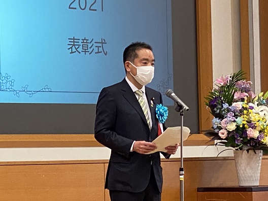 写真:マイクの前に立ち、挨拶をする井上大臣。胸には青い花の記章を着けている。