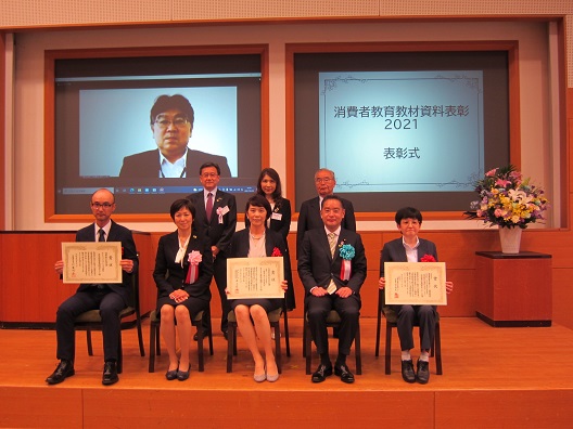 写真:受賞団体のメンバー7人と井上大臣が前後に並び記念撮影をしている。全列5人の右から2番目に井上大臣が座っている。8人の後ろには、大きなモニターが二つ映し出され、右側のモニターには「消費者教育教材資料表彰2021表彰式」左側のモニターには、1人の男性が映し出されている。