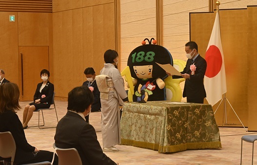 写真:受賞者である着物姿の女性に、表彰状を渡す井上大臣。大臣の横には、消費者ホットライン188のイメージキャラクターイヤヤンの着ぐるみが立っている。