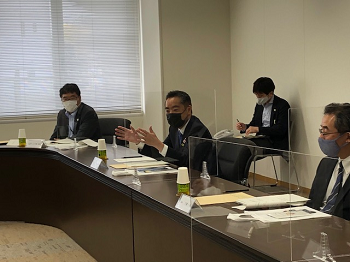 写真:繋げられた会議机の席に付き、発言している井上大臣。設置された席は一人づつ透明のパーテーションで区切られている。