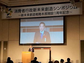 写真:消費者行政新未来創造シンポジウムの看板がかかる壇上の演台に立ち、開会挨拶を行う井上大臣。演台の後ろには、大きなモニターが設置され、挨拶する井上大臣の姿が映っている。