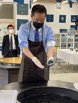 写真:黒いエプロンに、白のゴム手袋を装着し、藍染めの染料の入った入れ物に向かっている井上大臣。