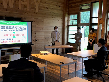 写真:取組説明のスライドが表示されたモニターの前の席に付き、説明を受けている井上大臣。