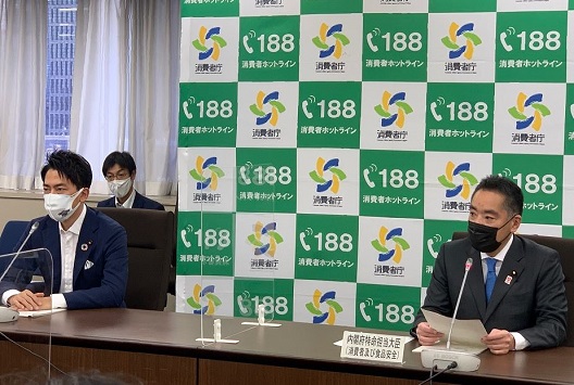 写真:消費者庁のロゴマークの並んだバックスクリーンの前に座り、発言している井上大臣。向かって左側には、パーテーションを挟み、小泉環境大臣が座っている。