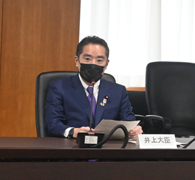 写真:着席し、発言をする井上大臣。黒いマスクをし、手に書類を持っている。机の上には、「井上大臣」の名札が置かれている。