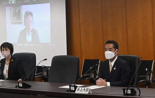 写真:写真中央、横に並べられた会議机に、着席し、マイクを前に話をする井上大臣。