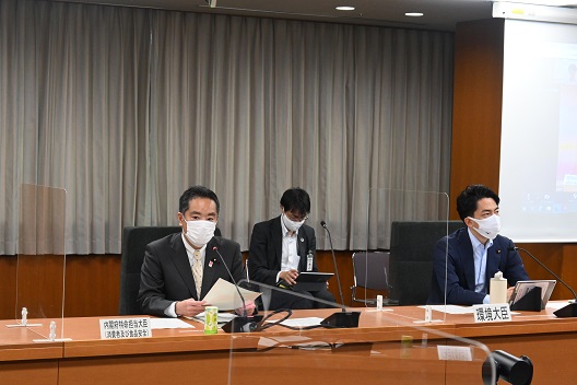 写真:写真中央、横に並べられた会議机に、着席し、マイクを前に話をする井上大臣。