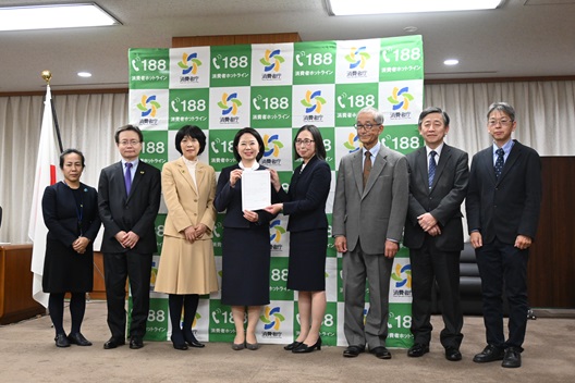 写真:消費者庁のロゴマークを背景に、8人の人物が並ぶ。中央で自見大臣と特定非営利活動法人消費者スマイル基金の代表が、認定証を掲げている