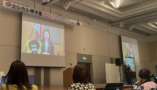 写真:エシカル甲子園2023の横断幕が掲げられた壇上で、スクリーンに自見大臣のビデオメッセージが放映されている。