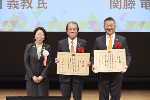 写真:自見大臣と受賞者2人が並ぶ。受賞者は、それぞれ表彰状をてに掲げている。