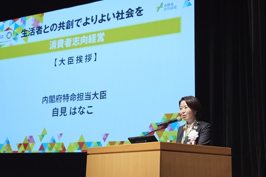 写真:演台にたち、話をする自見大臣。後ろのスクリーンには、「生活者との共創でよりよい社会を:消費者志向経営」の文字が表示されている