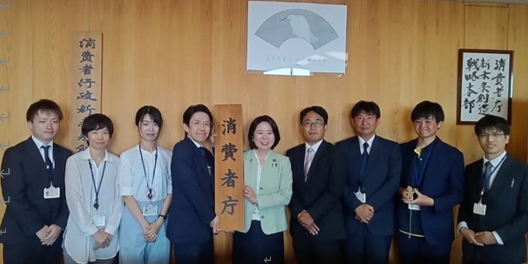 写真:「消費者庁」と書かれた看板を掲げた自見大臣を中心に、未来本部の職員が並んでいる