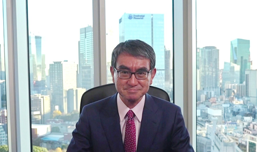 写真:窓を背景に、座っている河野大臣。窓の外には高層ビルの立ち並ぶ街並みが見える。