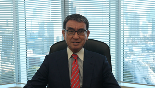 写真:窓を背景に、席についている河野大臣。後ろの窓には、高層ビルが並ぶ。