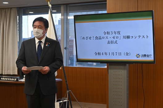 写真:モニターの横に立つ若宮大臣。モニターには「令和3年度「目指せ!食品ロス・ゼロ」川柳コンテスト表彰式」の文字が表示されている