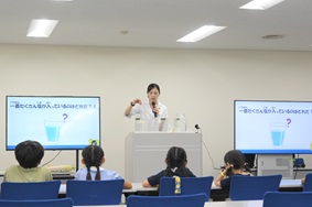 写真:白衣を着た講師が、参加者である子どもたちに、モニターで動画を見せながら説明している様子