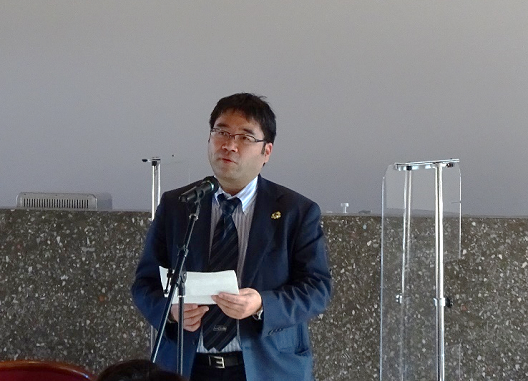 写真:スタンドマイクの前に立ち、話をするスーツ姿の男性