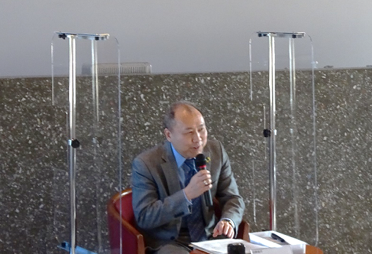 写真:ハンドマイクを手に持ち、座って話をするスーツ姿の男性