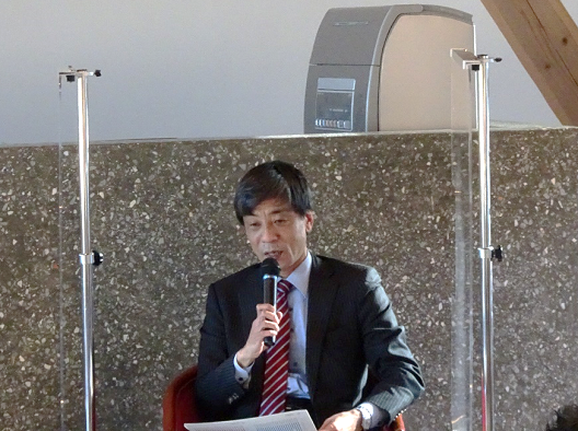 写真:ハンドマイクを手に持ち、座って話をするスーツ姿の男性