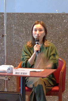 写真:一人掛けのソファと机のセットに座り、座って話をする女性。深緑のワンピースを着て、手にはハンドマイクを持っている。机の上には、鎌田安里紗氏の名札が置いてある。