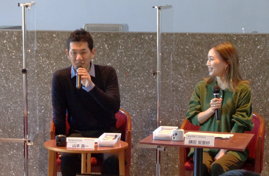 写真:一人掛けのソファと机のセットが二組並び、座る男女。二人の間は透明のパーテーションで仕切られている。左側に座っている男性がハンドマイクを手に持ち、話をしている。机の上には、山本 昌一氏の名札が置いてある。
