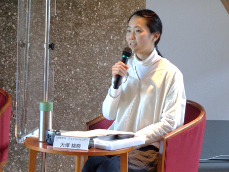 写真:一人掛けのソファと机のセットに座り、座って話をする白い服を着た女性。手にはハンドマイクを持っている。机の上には、大塚 桃奈氏の名札が置いてある。
